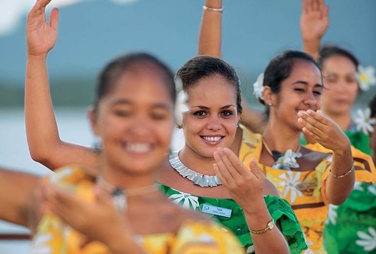 Dressed in alternating gold and green tropical print dresses, four members of Les Gauguines smile while performing a traditional polynesian dance. 