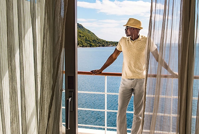 A man in a fedora enjoys the view and fresh air on his stateroom balcony.